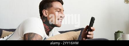 Handsome young man with tattoos smiles while checking his smartphone at home. Stock Photo