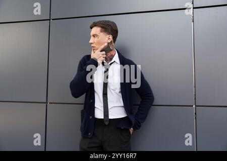 Tattooed man in a black cardigan and tie stands pensively by an urban backdrop. Stock Photo