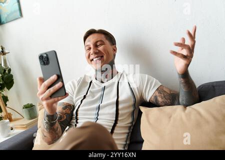 Young man with tattoos smiles joyfully while using his smartphone at home. Stock Photo