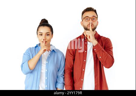 Focused young man and woman signaling quiet with fingers on lips, emphasizing silence or secrecy, dressed casually, looking thoughtful and serious, standing on an isolated white background Stock Photo