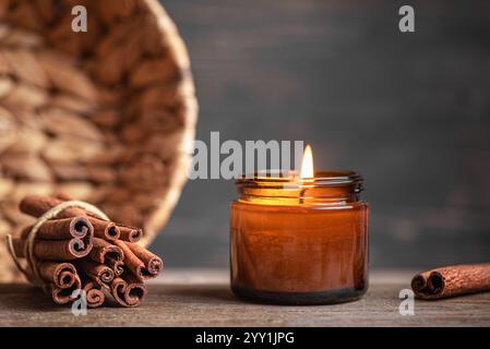 Scented burning candle in jar with cinnamon sticks on wooden table, copy sapce. Cosy hygge home interior decor, aroma massage with soy wax candle. Stock Photo