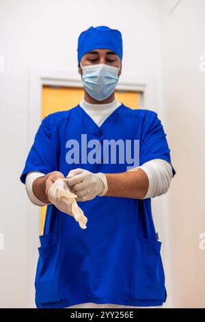 Young male dentist wearing blue surgical uniform, cap, and mask putting on sterile latex gloves, preparing for surgery inside dental clinic Stock Photo