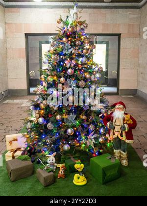 Christmas tree decorated with tinsel, garlands and balls with figure of Santa Claus and boxes with gifts in office hall Stock Photo