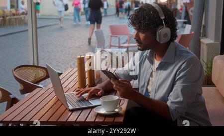 Indian Arabian man male businessman client business guy online work in cafe distant education using devices laptop phone listen music in headphones Stock Photo
