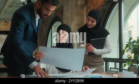 Muslim designer architect in hijab working in office drawing blueprints architecture project on desk business man women multiethnic team colleagues Stock Photo