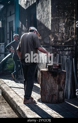 Traditional blacksmith at work in Alexandria, Egypt. A man hammers metal on an anvil atop a tree stump, surrounded by tools and sunlight-dappled walls Stock Photo
