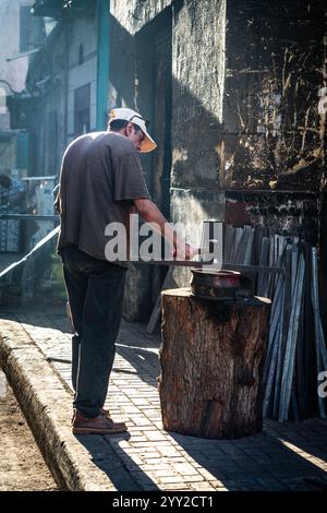 Traditional blacksmith at work in Alexandria, Egypt. A man hammers metal on an anvil atop a tree stump, surrounded by tools and sunlight-dappled walls Stock Photo