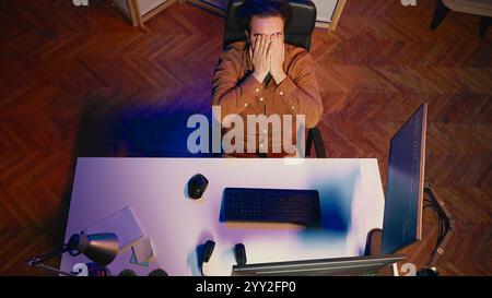 Top down view of software engineer putting face in hands in frustration while updating binary code script on computer, receiving errors. IT worker exasperated by annoying bugs while programming, aerial shot Stock Photo