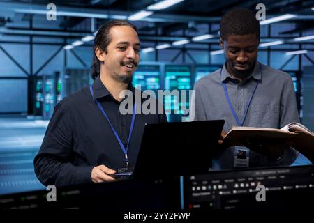 Cheerful IT specialists in server hub ensuring data remains shielded from potential threats. Smiling coworkers safeguarding data center against unauthorized access using laptop Stock Photo