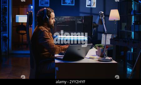 Developer focused on typing complex code in neon lit home office, listening music and developing software application. IT expert wearing headphones, solving coding project tasks on laptop, camera B Stock Photo