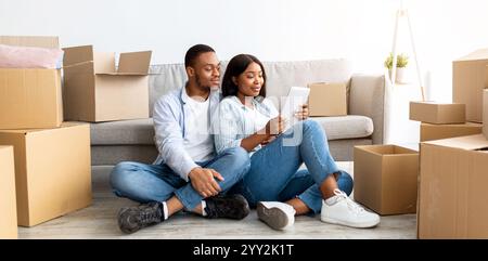 Loving african american spouses using tablet, searching new furniture while sitting among cardboard boxes, free space Stock Photo