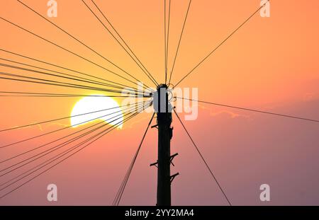 Silhouetted communications and power cables leading outwards  from a GPO telegraph pole with a large setting sun and orange sky   behind Stock Photo