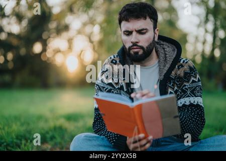A thoughtful man reads a book while sitting on grass in a tranquil park. Stock Photo