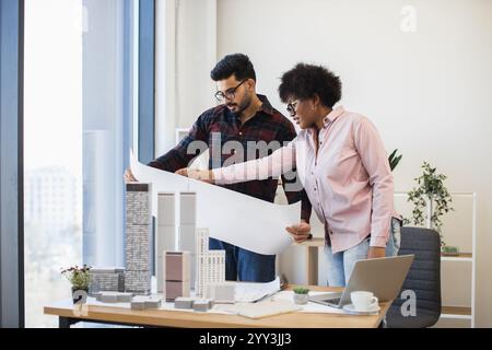 Indian man and African woman architects reviewing blueprints for developing modern city project. Collaboration and teamwork in architecture office setting. Stock Photo