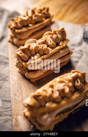Three rectangular mille feuille pastries topped with peanut butter cream and chopped peanuts are placed on a wooden serving board Stock Photo