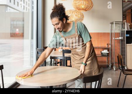 A cafe worker in an apron is attentively cleaning a table in a modern, cozy cafe setting Warm lighting and simple decor create a welcoming atmosphere, Stock Photo