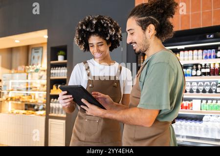 Two cafe workers in aprons smile while using a digital tablet, planning together in a vibrant cafe setting Shelves of drinks and snacks are visible in Stock Photo