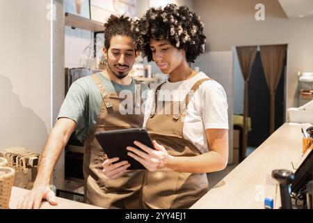 Two cafe workers in aprons are using a tablet behind a coffee shop counter, collaborating on tasks and planning The cozy ambiance highlights teamwork Stock Photo