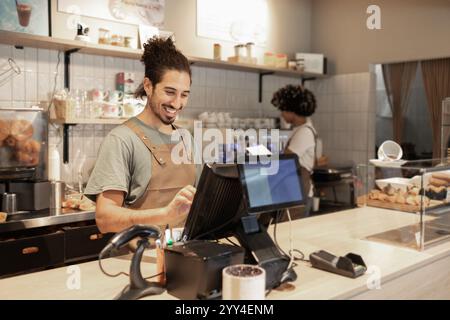 A cheerful cafe worker wearing an apron smiles while using the register at the counter Shelves with cups and pastries are visible in the cozy cafe set Stock Photo