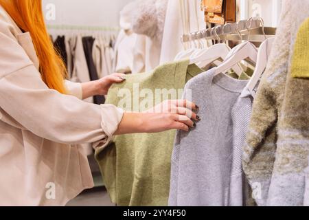 Cropped unrecognizable orange hair woman carefully selecting clothing items from a rack in a stylish fashion store. Her focus is on choosing the best Stock Photo