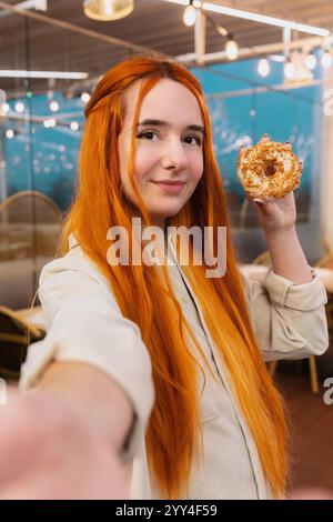 A young with orange hair woman is taking a selfie with her mobile phone while holding and eating a donuts in a cozy cafe. She looks directly at the ca Stock Photo