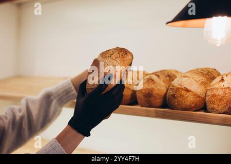 A gloved hand selects a fresh loaf of bread from a wooden shelf in a cozy bakery setting The artisanal loaves display a rustic, crunchy crust under wa Stock Photo