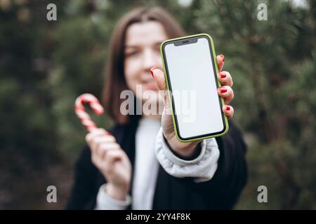 A blurred woman wearing a cozy outfit holds a smartphone with a blank screen and a classic red and white candy cane, symbolizing the Christmas season Stock Photo