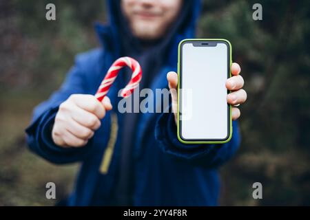 A person in a blue coat holds a classic red and white candy cane in one hand and a smartphone with a blank screen in the other, set against a natural Stock Photo