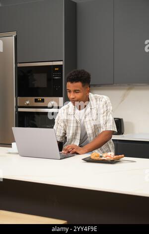 A young Black man is focused on his laptop in a modern kitchen, capturing a moment of productivity and comfort The atmosphere is relaxed, with pastrie Stock Photo
