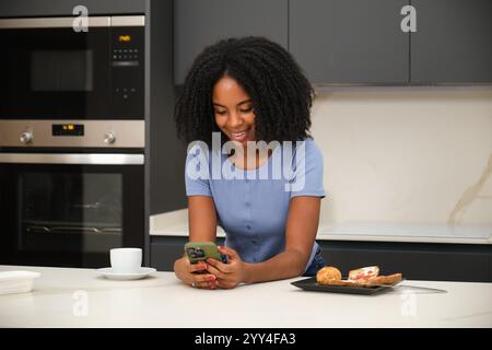 A young African American woman shares a joyful moment at home The woman smiles while using a smartphone in the kitchen The scene captures warmth and e Stock Photo