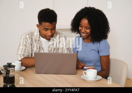 A young Black couple sitting at a table, browsing and shopping online using a laptop and credit card They are smiling and enjoying coffee together in Stock Photo
