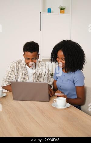 A cheerful young black couple sits at a wooden table in their modern home, engaged with a laptop and drinking coffee The atmosphere is relaxed and joy Stock Photo