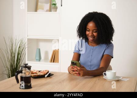 A young African American woman enjoys coffee while using her smartphone at a cozy home setting, blending relaxation with modern technology for a casua Stock Photo