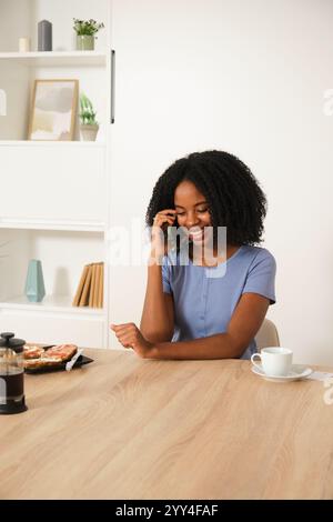 A young African American woman smiling while talking on the phone at home She's seated at a wooden table with breakfast and a cup of coffee, creating Stock Photo