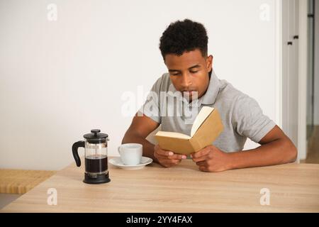 An african american man is sitting at a wooden table, engrossed in a book A French press and a cup of coffee suggest a cozy and relaxing home environm Stock Photo