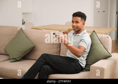 An African American man enjoys a calm evening at home He is seated comfortably on the couch, holding a remote control, creating a cozy and relaxed atm Stock Photo