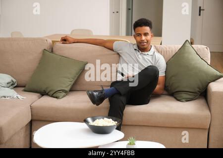 A young Black man relaxes on a comfortable beige sofa in a stylish, modern living room He holds a remote control, with popcorn on the table and a calm Stock Photo