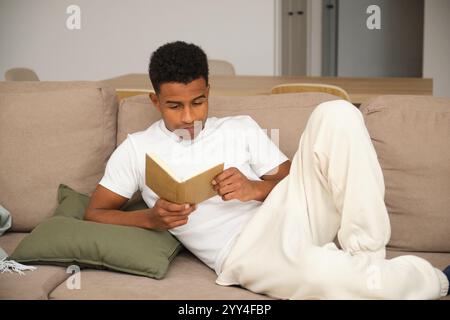 A young African American man relaxes on a beige sofa, reading a book The atmosphere is cozy and tranquil, emphasizing leisure and comfort in a modern Stock Photo