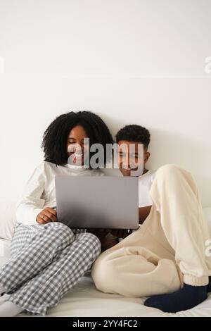 A young Black couple sits comfortably on their bed, sharing a laptop They are in casual pajamas, enjoying a relaxed moment together at home, smiling a Stock Photo