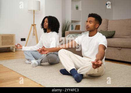 A young black couple sits cross-legged on the living room floor with eyes closed, practicing mindfulness meditation The cozy home backdrop adds warmth Stock Photo
