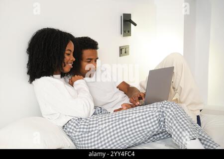 Young black couple lounging comfortably on a bed, each dressed in casual attire, enjoying a moment together while using a laptop in a cozy home settin Stock Photo