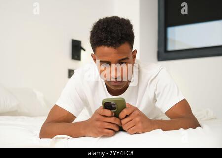 A young African American man is seen relaxing at home, concentrating on his smartphone while lying on a comfortable bed in a modern, well-lit bedroom Stock Photo