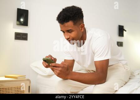 A young African American man sits comfortably on a bed, engrossed in his smartphone The cozy home setting reflects a relaxed and intimate atmosphere Stock Photo