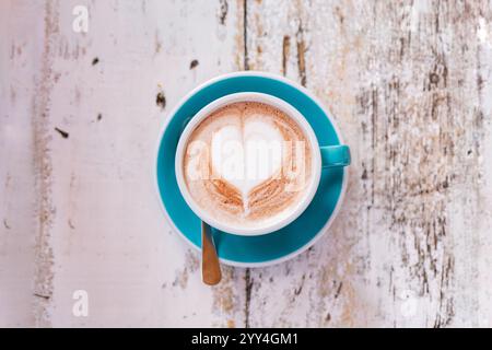 A cappuccino in a turquoise cup features a heart-shaped latte art design. The cup rests on a rustic white wooden table, creating a cozy and artistic a Stock Photo