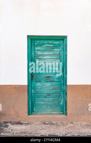 A charming teal wooden door set against a rustic white wall with a tan base. The door's weathered appearance adds character to the simplistic architec Stock Photo