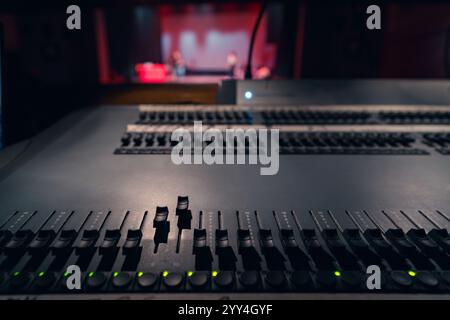 A close-up view of an audio mixing console in a dimly lit theater setting. The stage in the background is blurred, with figures performing under soft Stock Photo