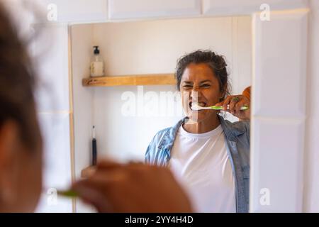 A woman brushes her teeth, standing in front of a bathroom mirror. Her reflection shows focus on dental hygiene, wearing casual attire with a bright b Stock Photo