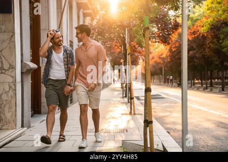 A gay couple enjoys a romantic walk on a tree-lined city street bathed in warm sunset light, showcasing affection and joy in their companionship Stock Photo