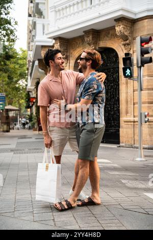 A joyful gay couple shares a tender moment, hugging and smiling on a bustling city street, carrying a shopping bag and enjoying a sunny day Stock Photo