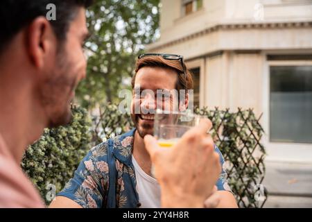 A gay couple shares a joyful moment, sipping drinks and engaging in a light-hearted conversation on a city terrace Stock Photo